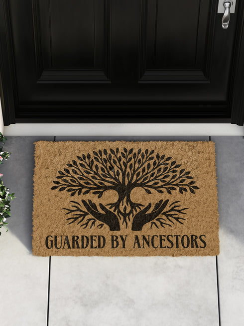 18x24 inch rectangular coir doormat made from natural coconut husk fibers, featuring a bold black print of a stylized tree with branching leaves and intertwined roots forming the shape of protective hands. Below the tree design, the phrase "GUARDED BY ANCESTORS" is printed in large, capitalized serif lettering. The mat has a textured, bristled surface ideal for scraping dirt and moisture from shoes, and a durable construction suited for everyday entryway use. Designed for indoor or outdoor placement, this d